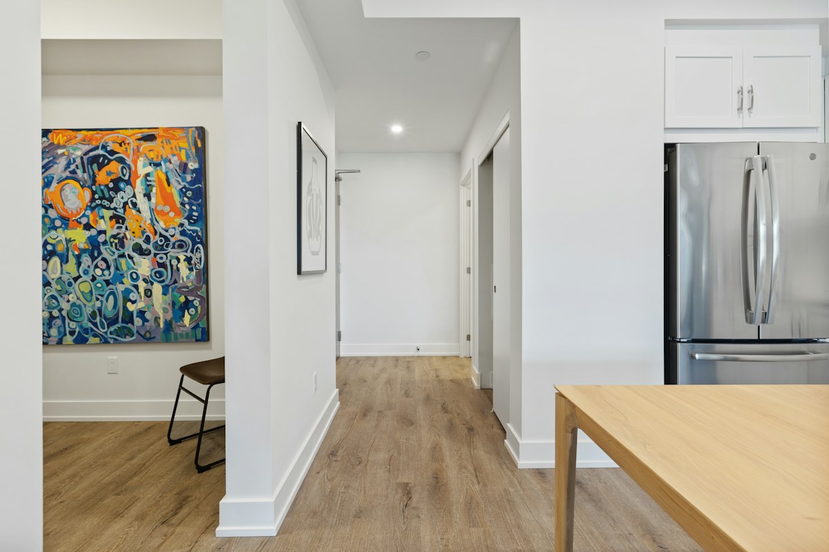A pale hallway adjoining a kitchen, with timber flooring and a colorful framed picture.