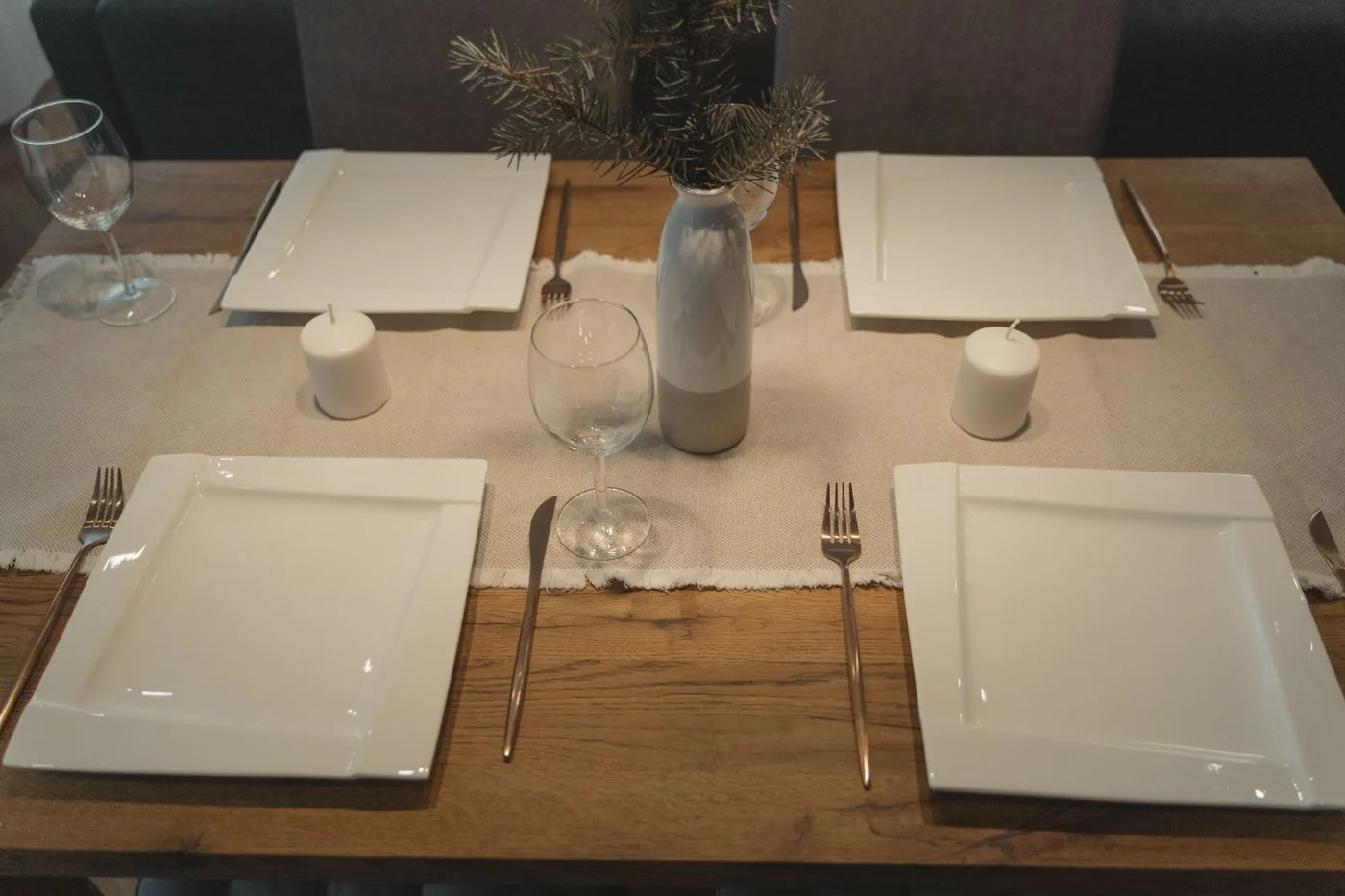 Square white plates, pale napkins and glasses on a wooden dining table.