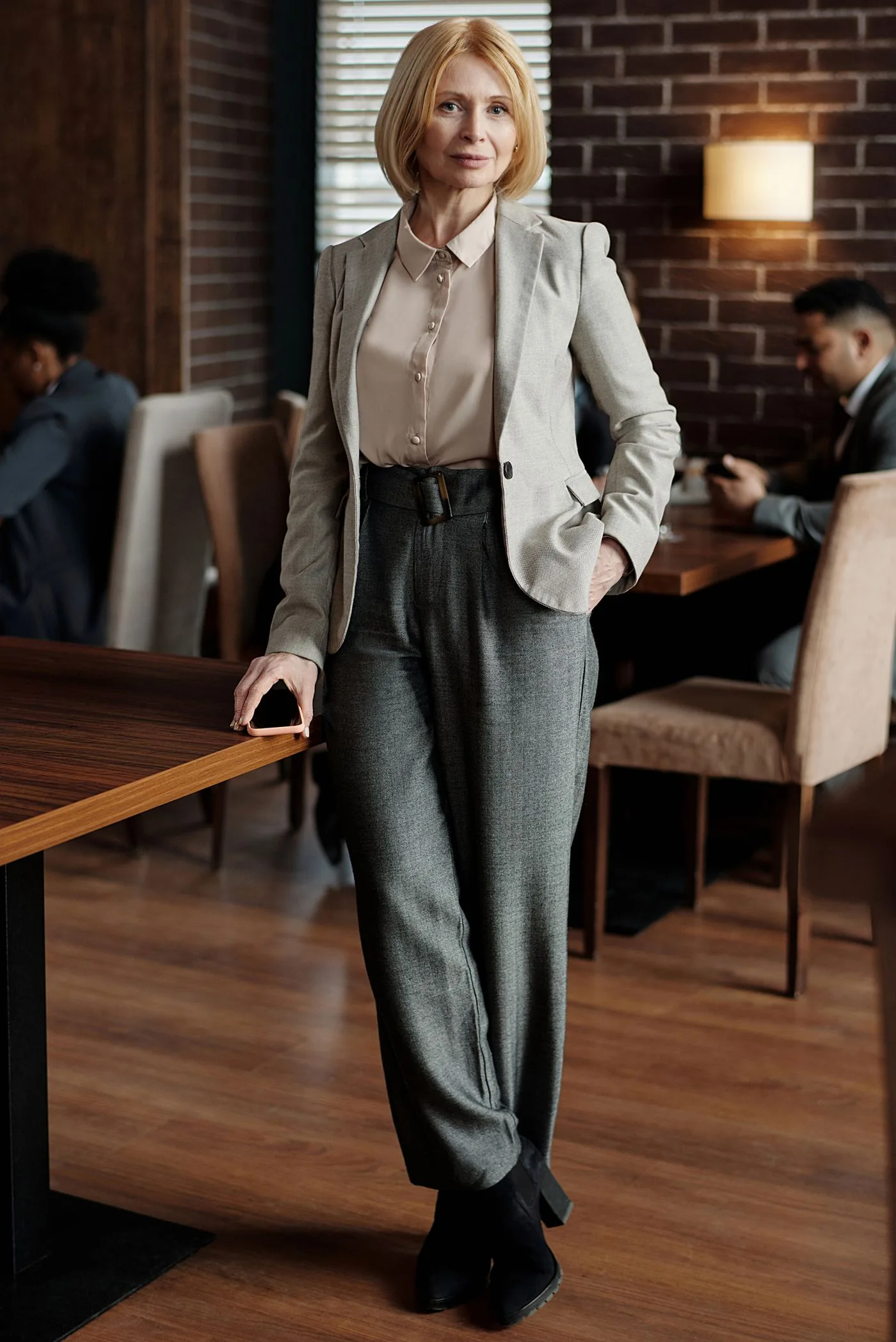 A woman in a beige blazer and gray trousers standing beside a café table.