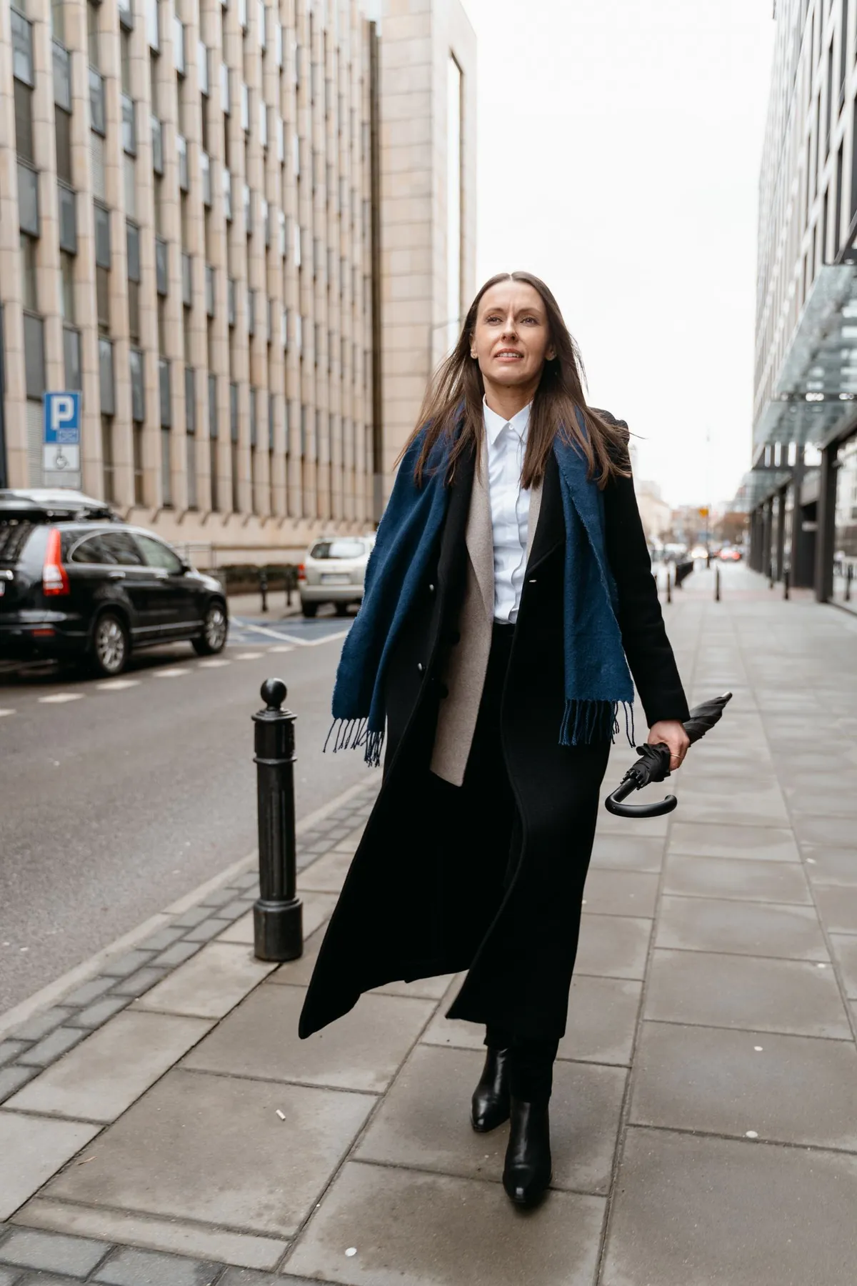 A woman wearing a dark coat and blue scarf carries a closed umbrella on a city pavement