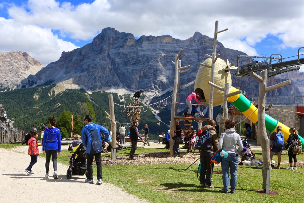 families with pushchairs, walking sticks, backpacks playing on a playground with climbing frame surrounded by mountains 