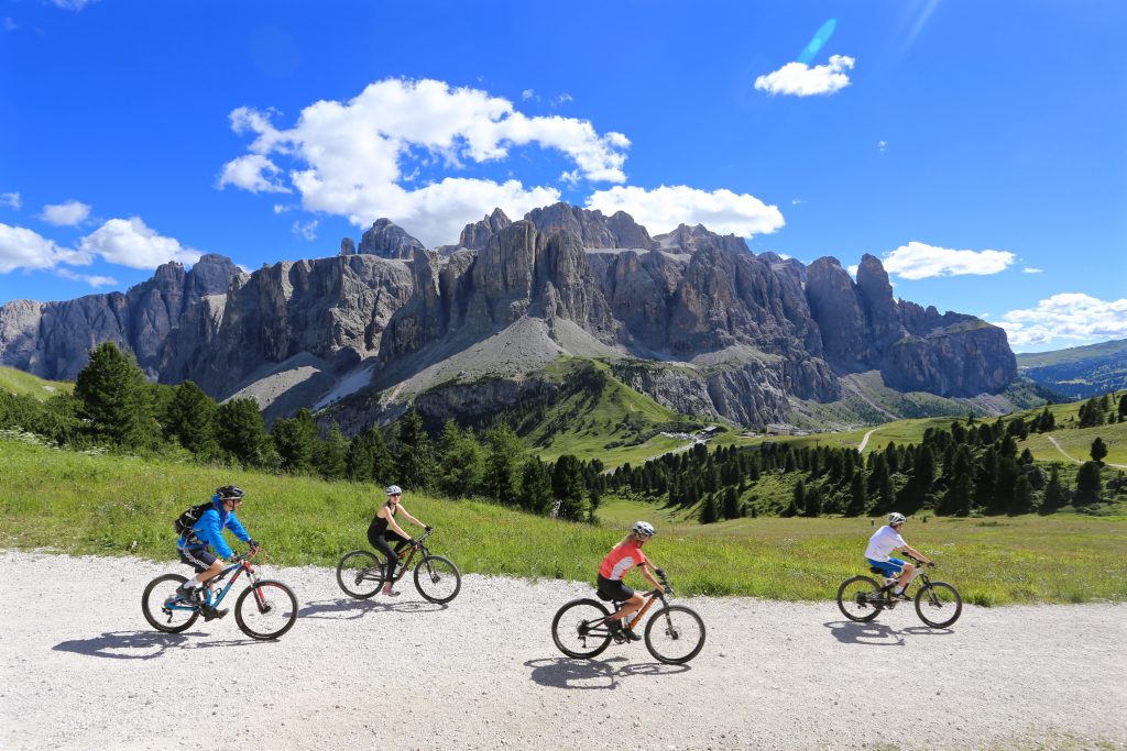 view of the mountains with 4 cyclists biking to the right on a gravelled path in the forefront of the picture