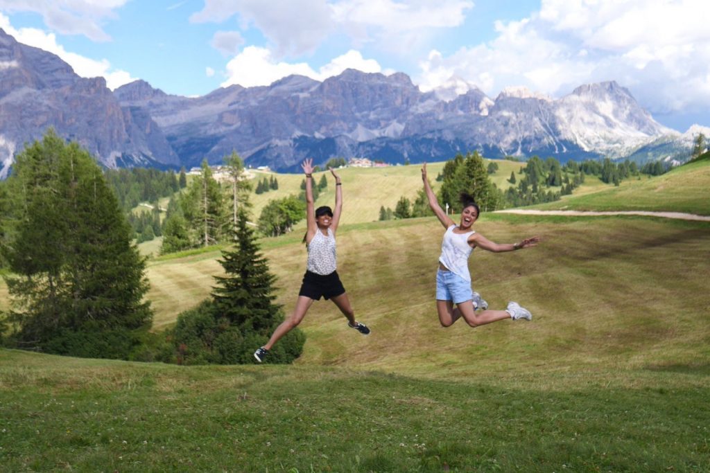 sima and friend jumping mid air with the dolomites in the background, grass, fields and mountains