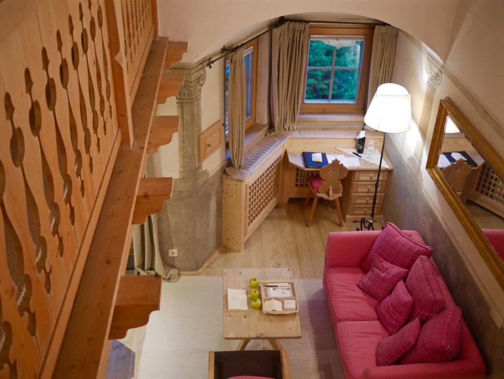 view from the balcony/mezzanine looking down to the living area with pink sofa, wooden furnishing and three apples on a table inside the Rosa Alpina suite