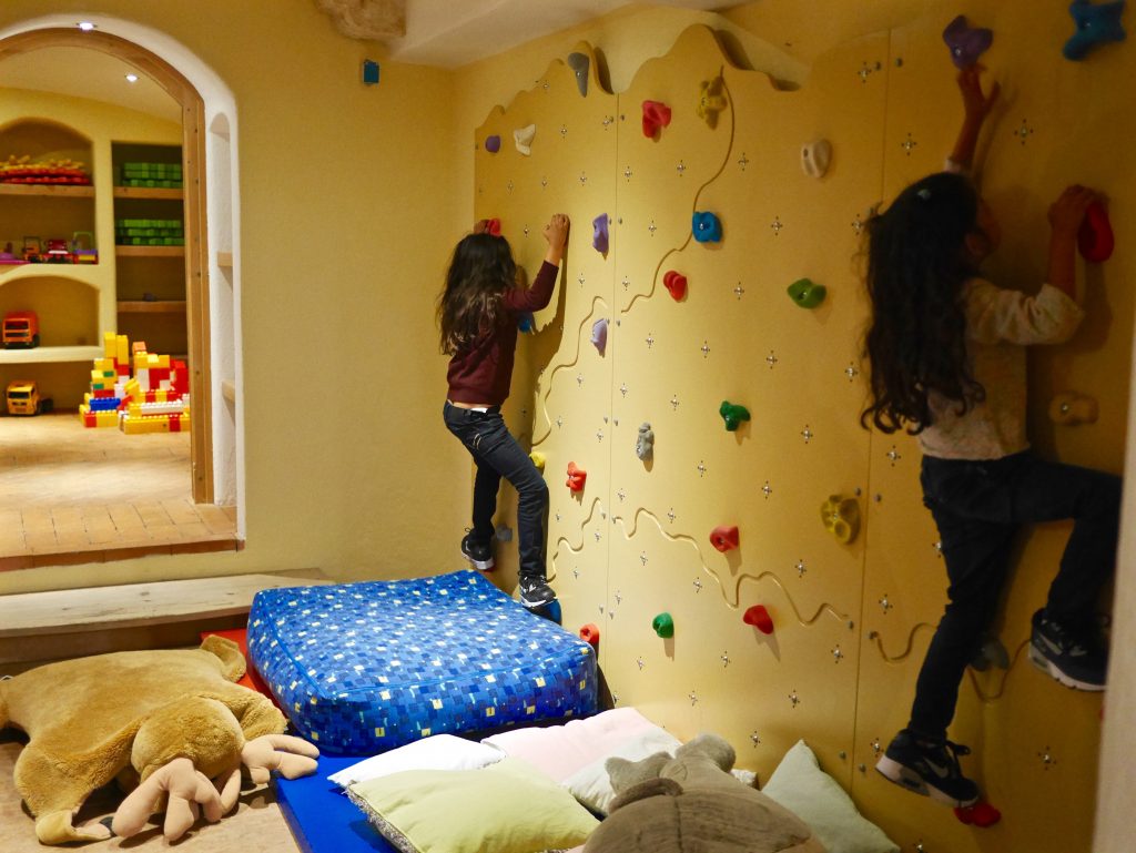 kids room with climbing wall surrounded by soft furnishing on the floor & two girls climbing the wall
