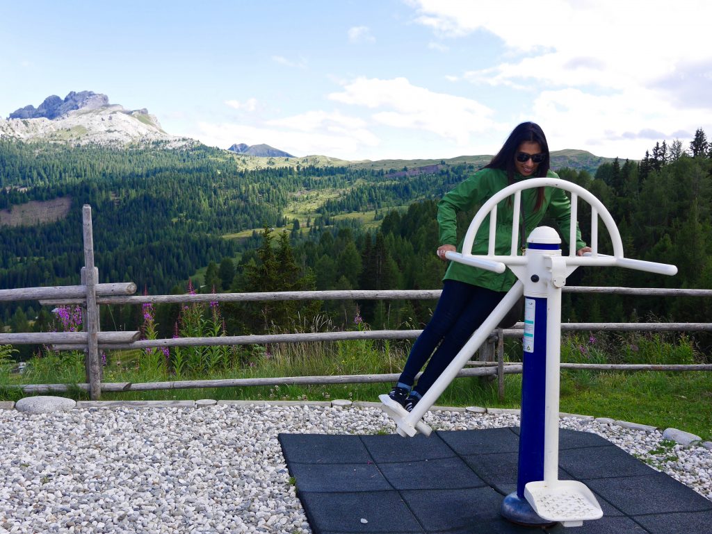 sima on a piece of fitness equipment in a green jacket and sunglasses overlooking the greenery and valleys of the dolomites
