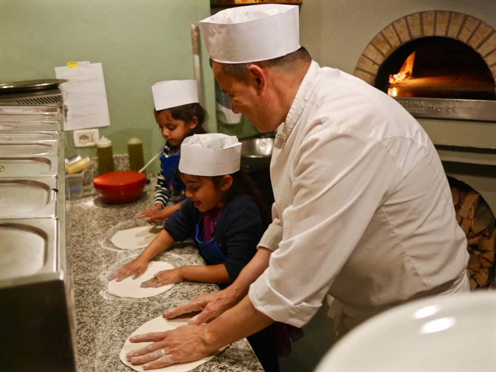 two children in white chef hats making pizza along side the chef with the pizza oven in the background