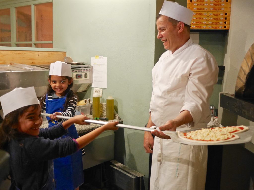 two girls in blue aprons and white hats putting their heart shaped pizza in the oven with help from the chef also in a white hat and kitchen wear