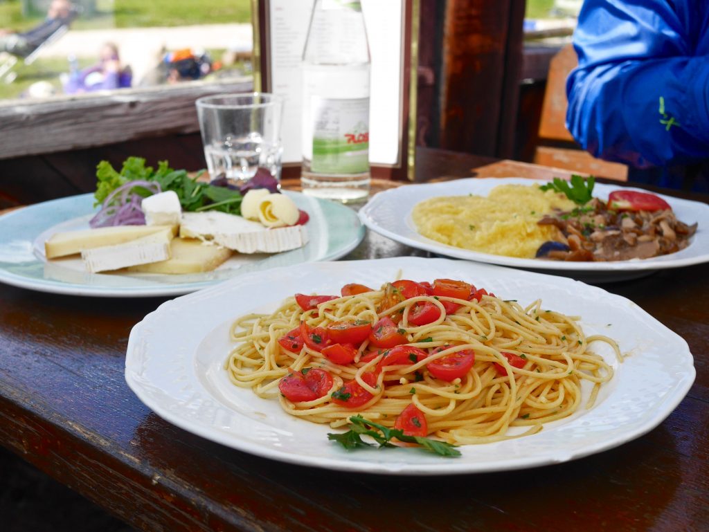three plates of food including one with tomatoes, herbs and spaghetti on a table along side a bottle of water and a glass.