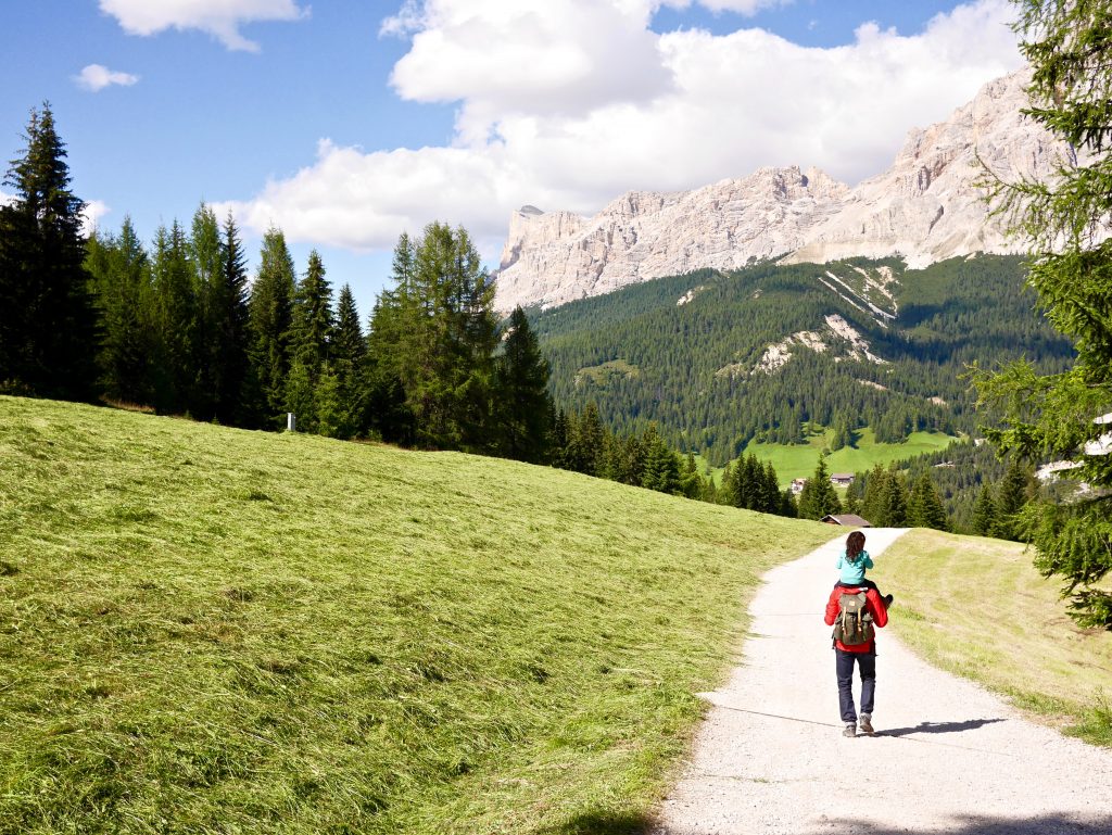 man in. red coat and brown back pack holding girl in blue jacket and black hair on his shoulders walking towards the mountains on a gravel path