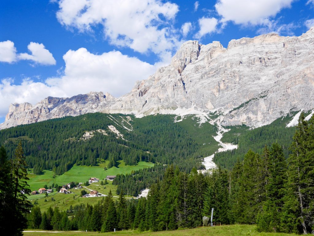 view of the Dolomites looking up the mountains. Lower half greenery and trees plus small buildings upper half snow/cloud covered mountains with blue skies