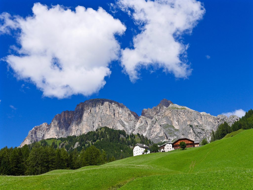 looking up towards the mountains surrounded by green grass, facing a white painted resort, wooden resort and blue skies
