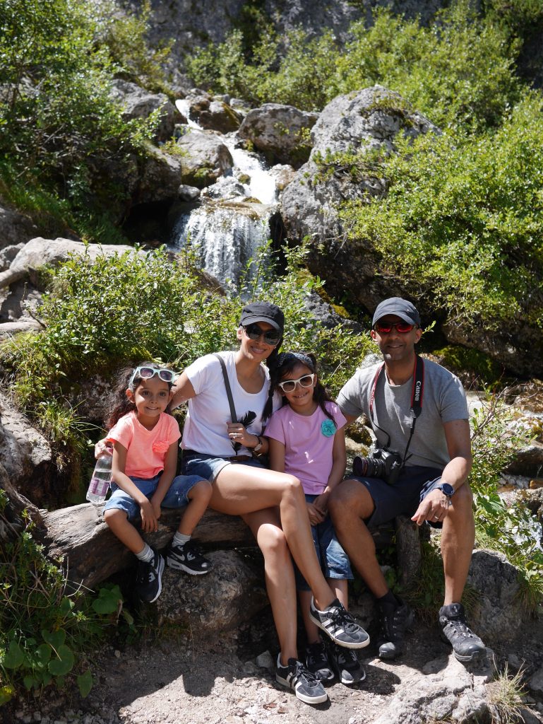 view of sima in white t-shirt and black cap and her family, girl in orange t-shirt, green glasses and denim short, girl in white sunglasses, purple t-shirt and denim short and husband in red reflective sunglasses, canon camera around his neck with grey t-shirt and blue shorts in front of the waterfall