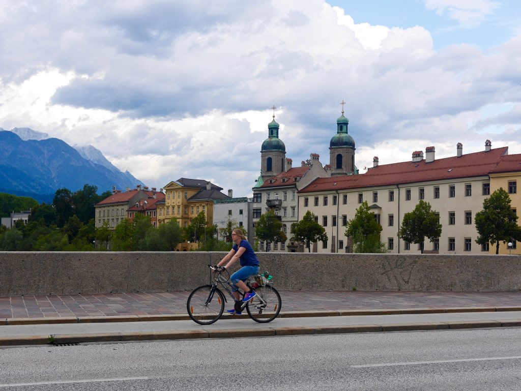 picre of a lady on a push bike closing a bridge surrounded by mountains and red tiled buildings with small square windows in Innsbruck