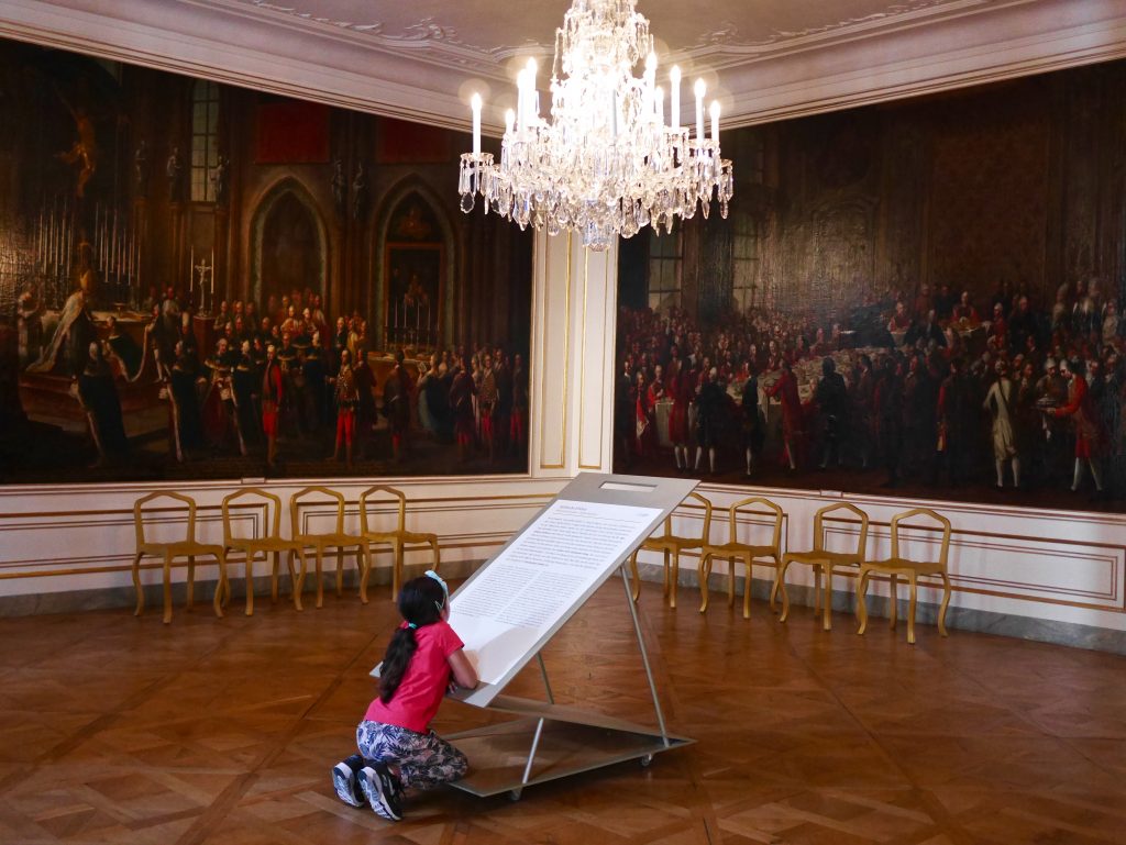child reading a display in the Hofburg palace under big crystal chandelier surrounded by painted walls and gold chairs