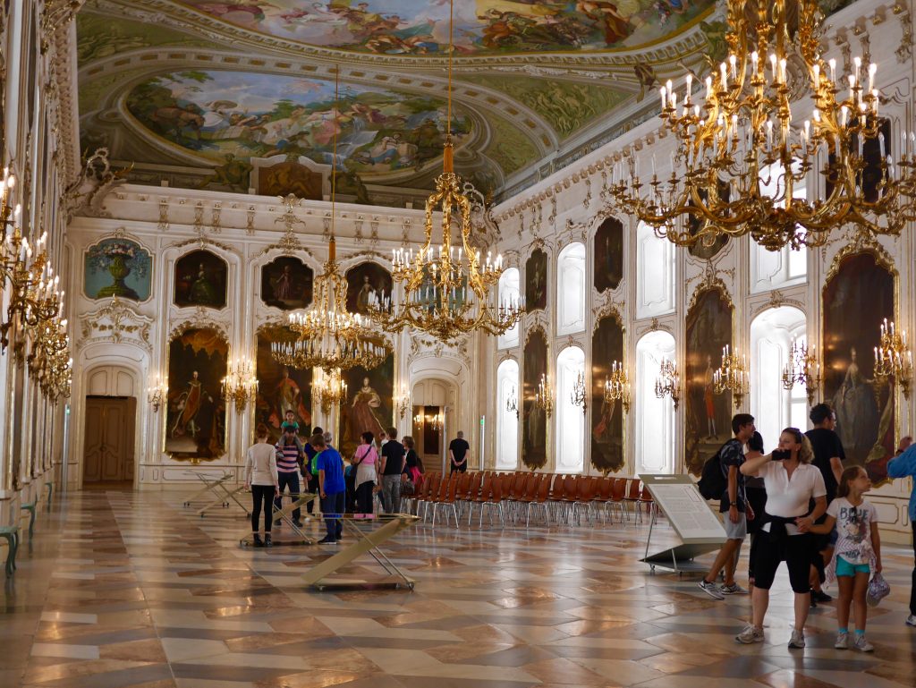 inside the giant hall of the Hofburg palace with white walls, gold architecture, gold chandeliers, painted ceiling and tiled floors