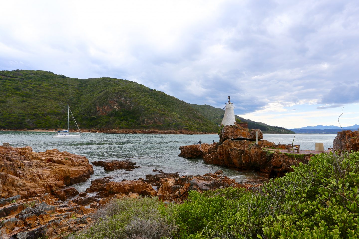picture looking out to the lagoon surrounded by greenery, brown rocks and a white yacht on the water