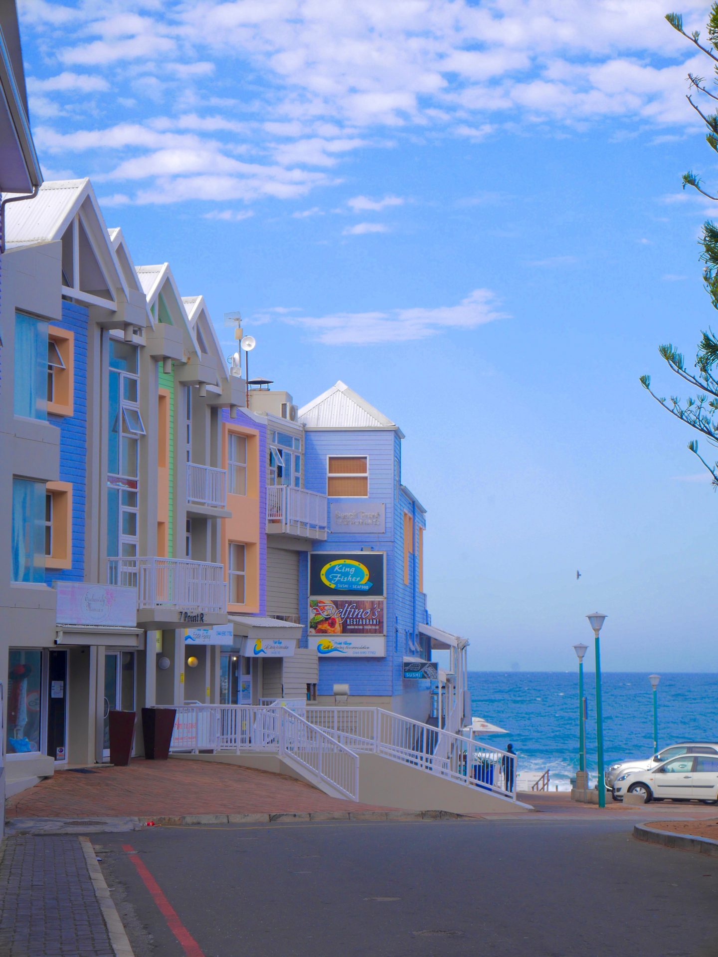 pastel coloured 2 story high buildings under blue skies looking down beach of Mossel bay
