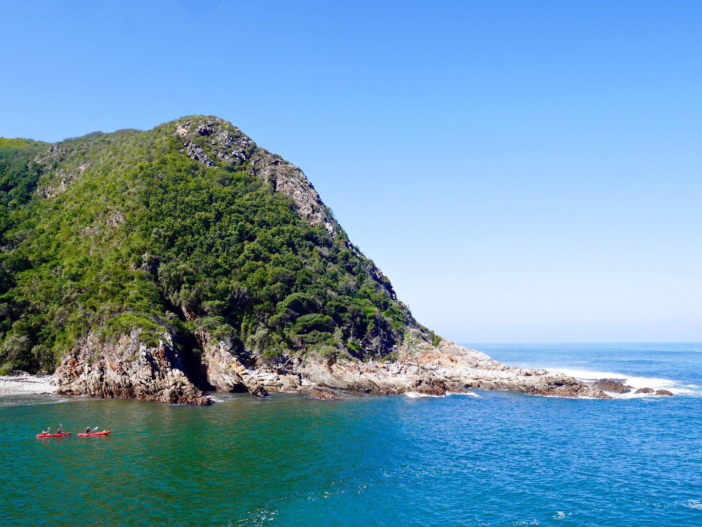 picture of a cliff on the edge of Port Elizabeth with green shrubbery, white cliffs and stone and two red canoes in the green blue water