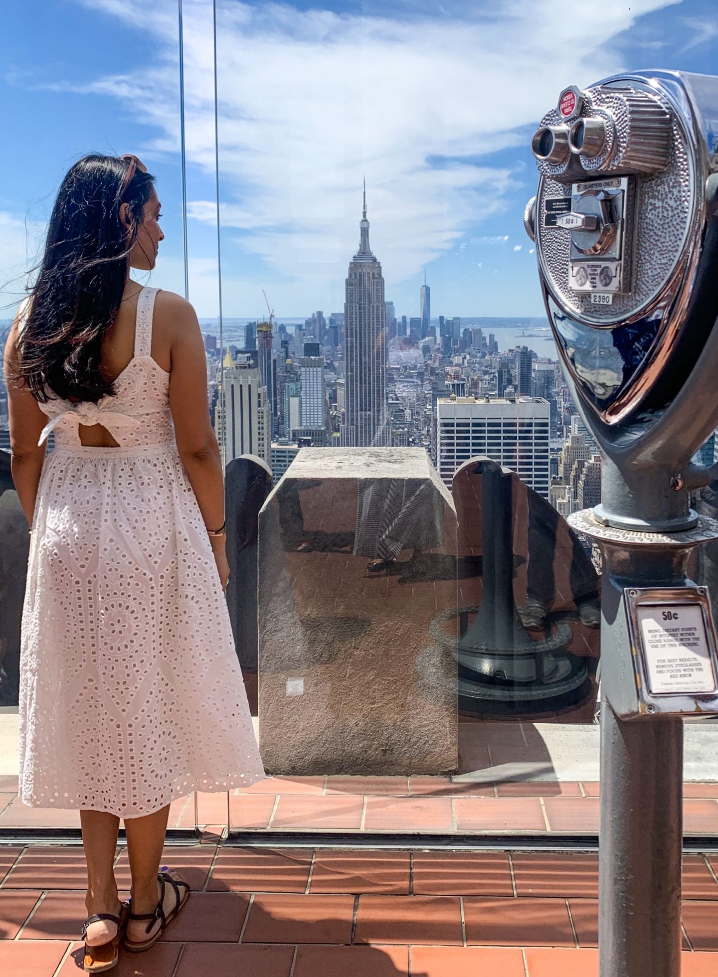 sima in white dress and sandals along side 50c viewing binocular admiring view of Empire State Building from Top of The Rock