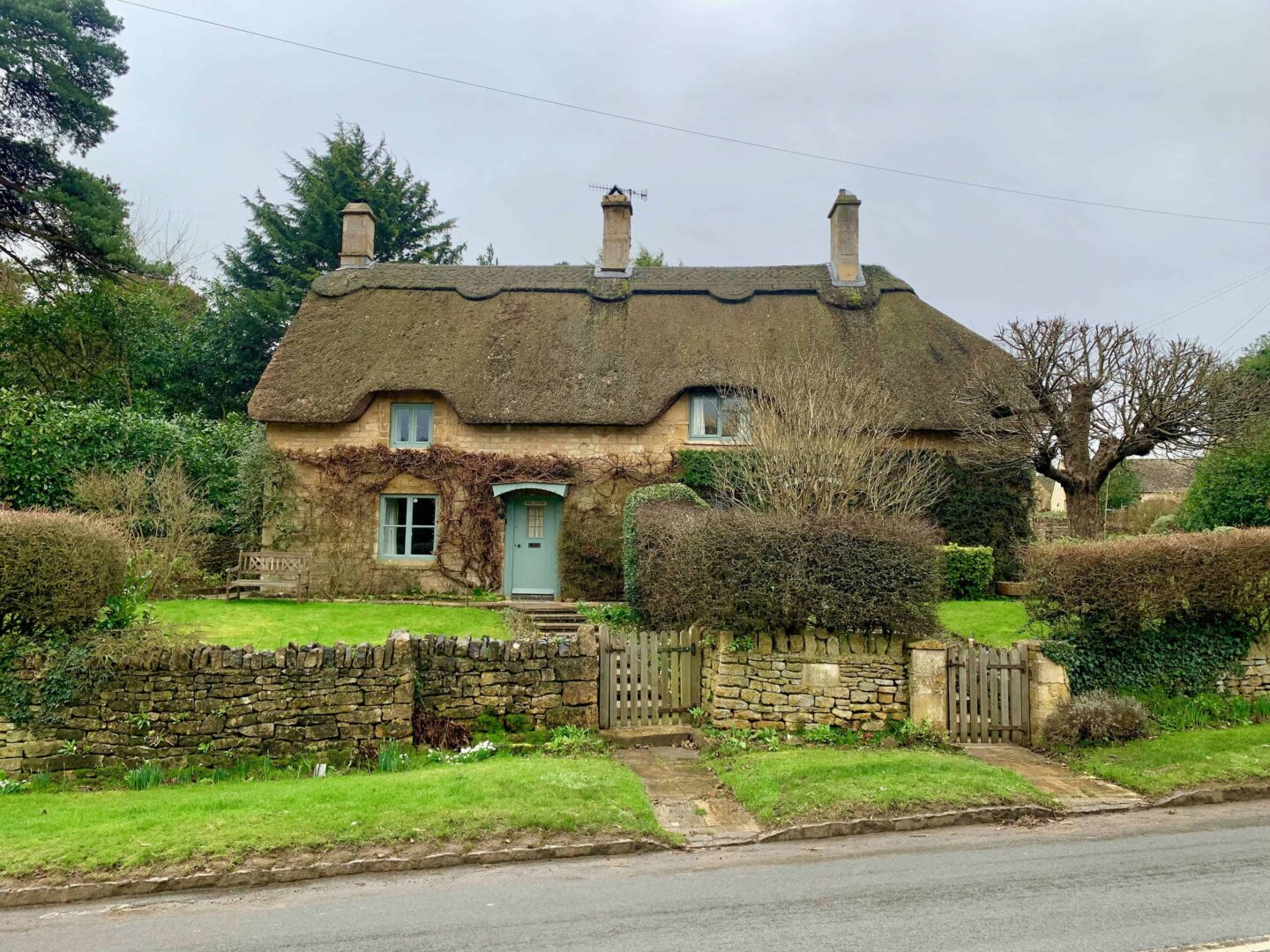 view of a thatched roof cottage with three chimney stacks, winces and green grass with a picket fence of a road in The Cotswolds