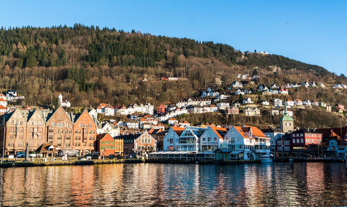 view from a lake in Norway looking towards the three+ story buildings and fir trees alongside the mountain