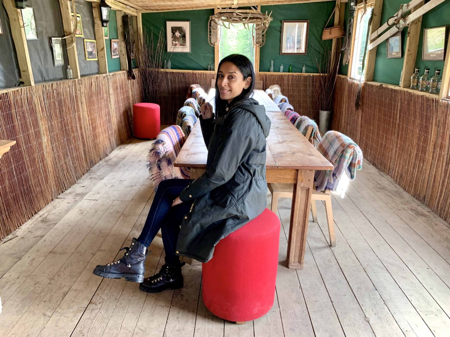 view of sima sat on a red stool inside the wooden cabin against a long wooden table with blankets covering the chairs