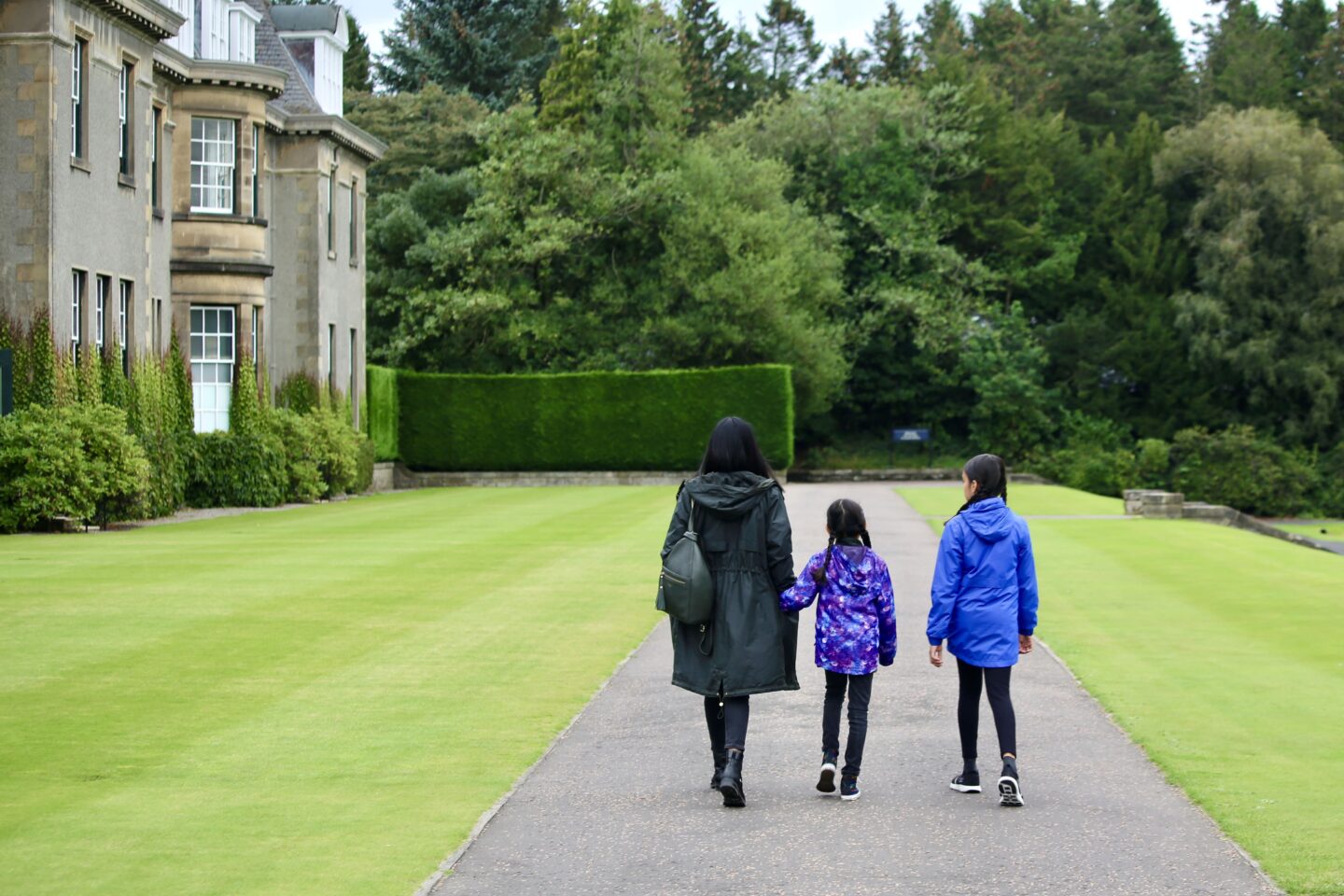 view of sima holding the hand of younger daughter in purple rain coat alongside eldest in blue walking along the path with gleneagles hotel to the right surrounded by grass