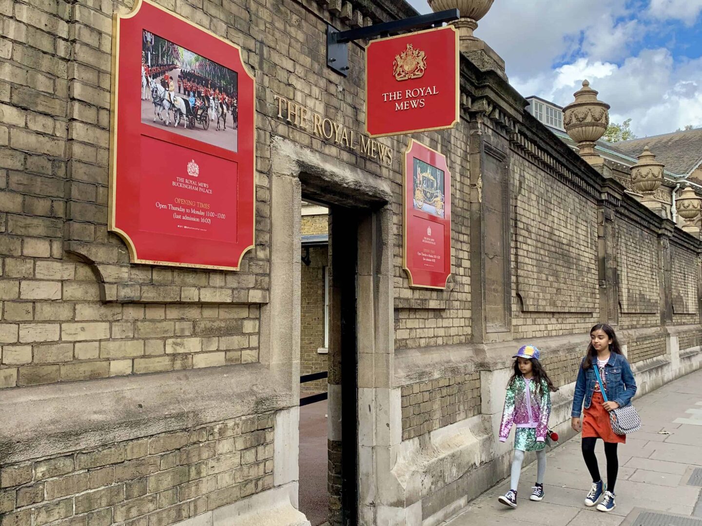 two brown haired girls holding handbags and wearing bright coloured clothing walking along the street towards the entrance of the royal mews